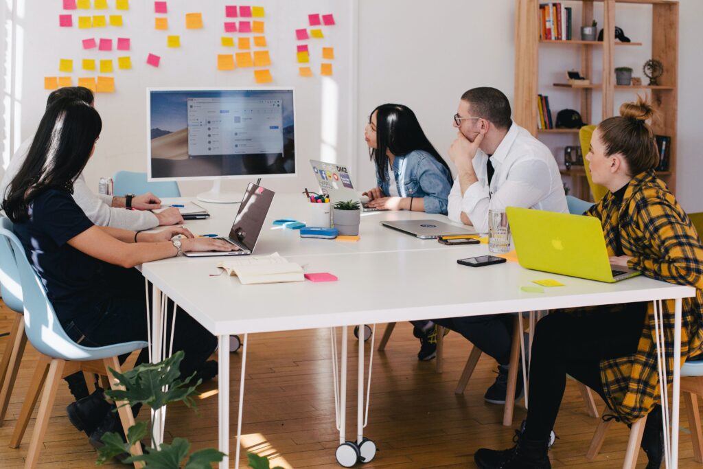 People sat around an office table. There are sticky notes and laptops. They are looking at a presentation on a screen.