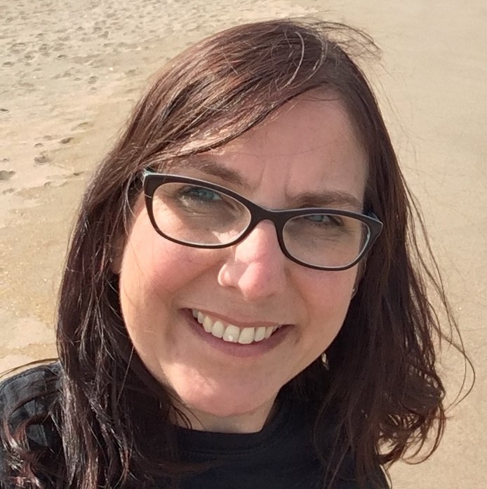 Helen Redman smiling on a beach, brown shoulder-length hair and glasses. 