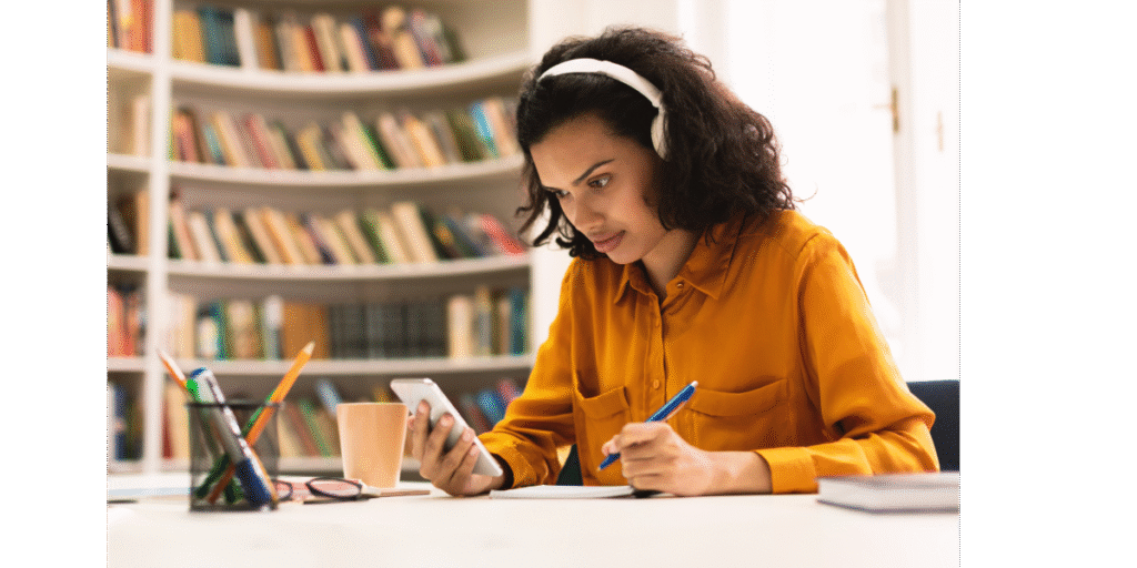 Female student wearing headphones and looking at her phone. She is in a library.